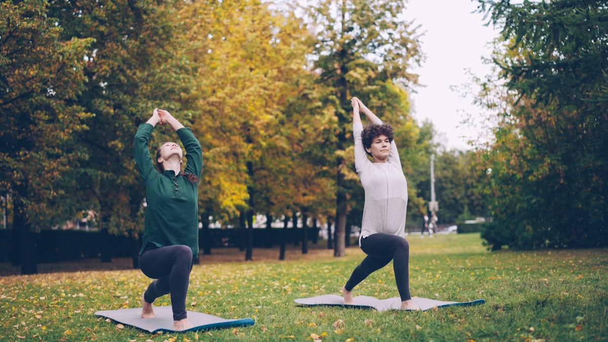 Slender girl yoga student is learning Eagle pose under guidance of experienced teacher during individual practice with instructor in park. Beautiful autumn nature is in background.